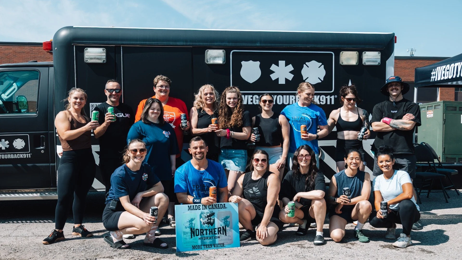 Group of people standing outside in front of a black ambulance with medical symbols. They are smiling, holding cans, and some are wearing athletic clothing. There is a sign that reads 'Made in Canada. Northern Hydration' in the front.