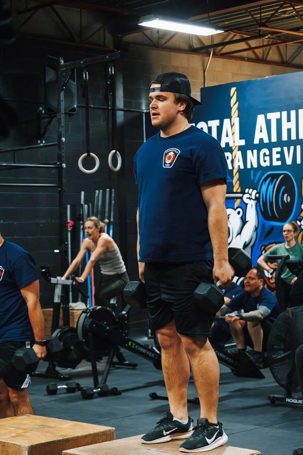 A man is lifting dumbbells during a workout at the gym, with other people exercising in the background. The gym has a blue banner with the text 'TOTAL ATHLETIC ORANGEVILLE' and a mural of a bear lifting weights.