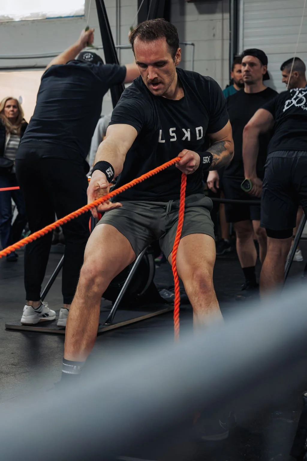 A man participating in a fitness competition or workout session, pulling a thick orange rope while seated on a chair, with a group of people watching around him.