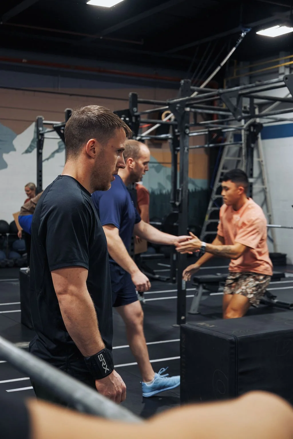 People at the gym, two men shake hands, others working out, gym equipment visible in background.