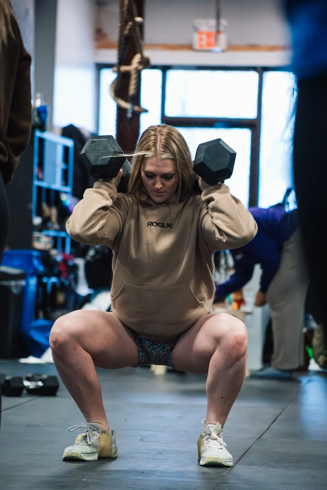 A woman in a beige hoodie and sneakers is squatting while lifting two black dumbbells on her shoulders, inside a gym.