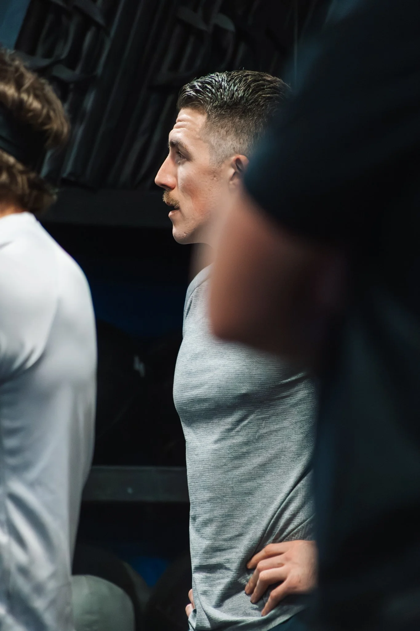 Side view of a man with slicked-back hair and a mustache, wearing a gray shirt, with hands on hips, in a dimly lit indoor setting.