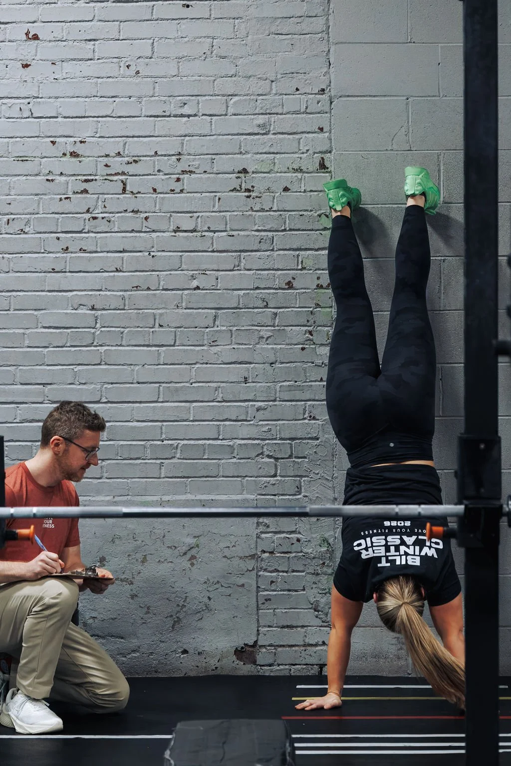 A woman in black leggings, green shoes, and a black 'Built Warrior Classic' shirt is doing a handstand against a gray brick wall, with her legs extended upward. A man in glasses, a reddish-brown shirt, and beige pants is kneeling beside her, taking n
