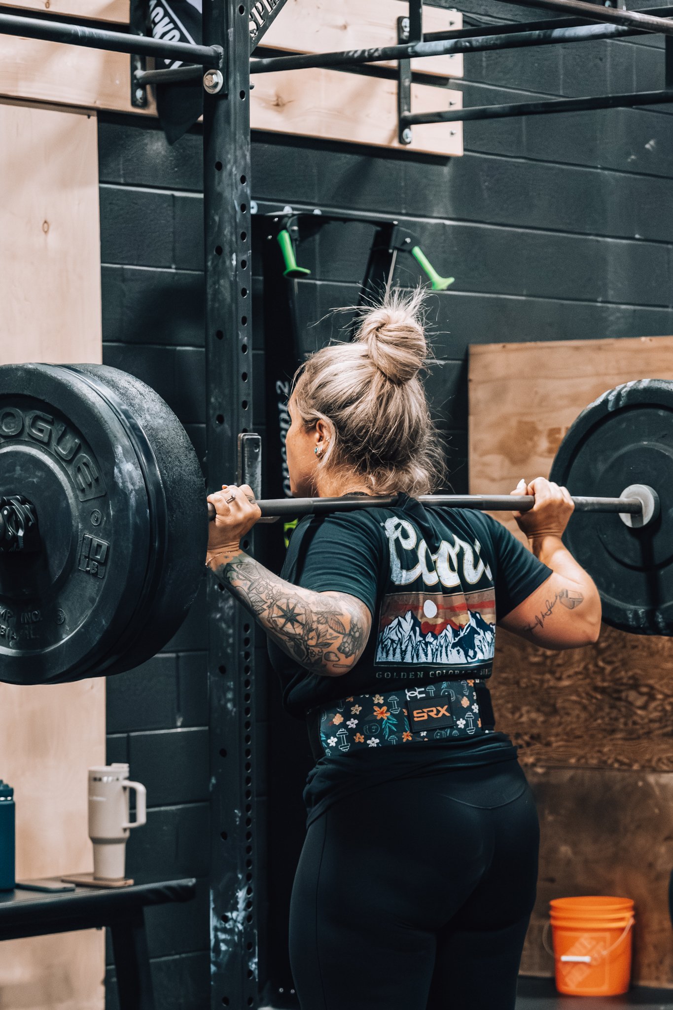 A woman with tattoos on her arm and bun hairstyle lifting a loaded barbell during weightlifting workout in gym.