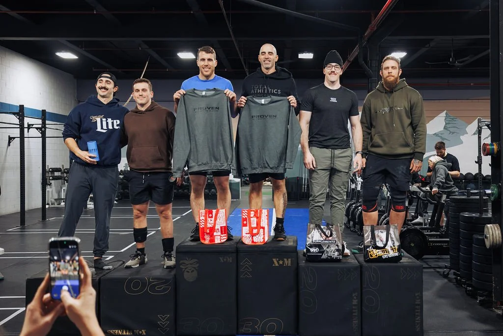 Six men standing on a podium inside a gym, holding prizes and apparel, with gym equipment in the background. One person in the foreground is taking a photo.
