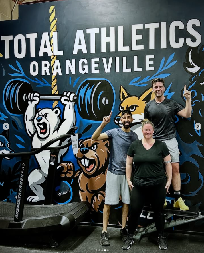 Three people posing in front of a colorful gym mural with animals lifting weights, including a polar bear, a lion, and a fox, inside Total Athletics Orangeville gym.