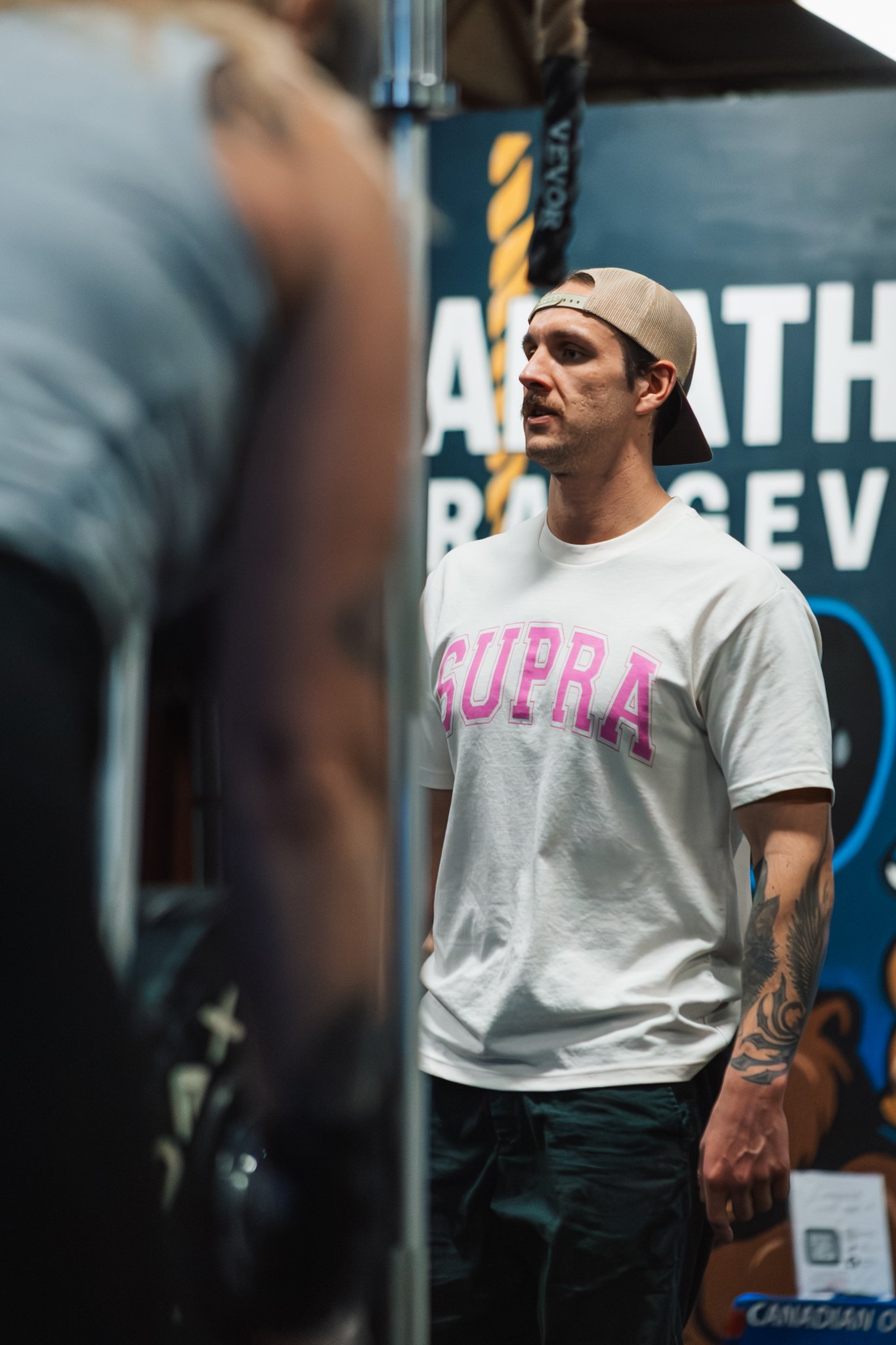 A man wearing a beige cap backwards, a white t-shirt with pink lettering, and tattoos on his left arm, standing in front of a workout gym banner.