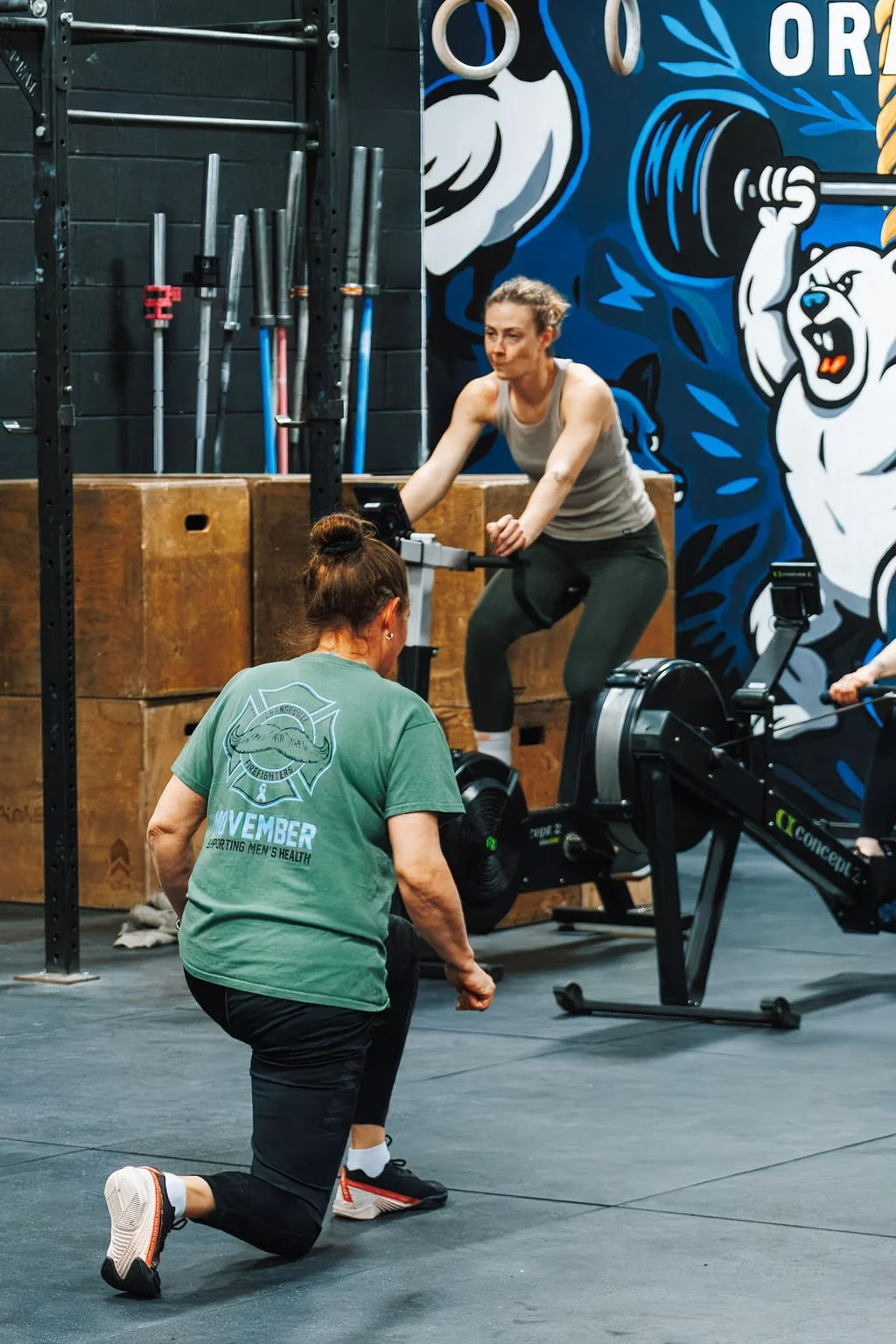 A woman working out on a rowing machine with a trainer instructing her in a gym, with a colorful mural of bears on the wall behind them.