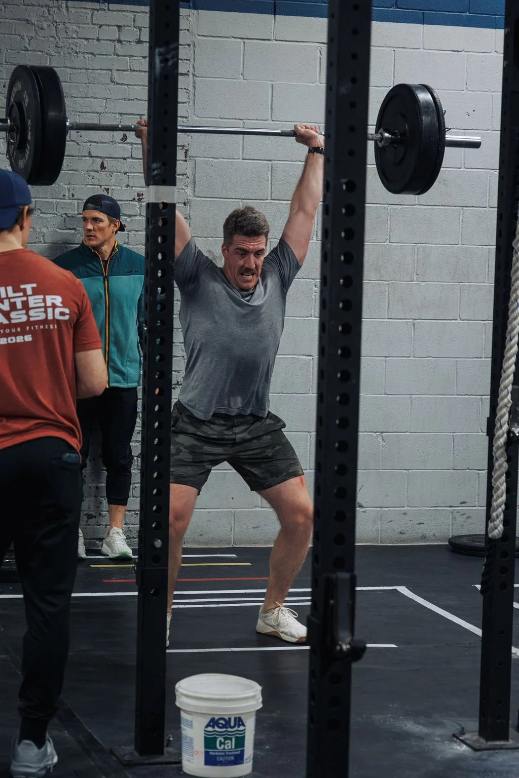 A man lifting a barbell with weights in a gym, surrounded by others observing, with a bucket of AquaCal on the floor.