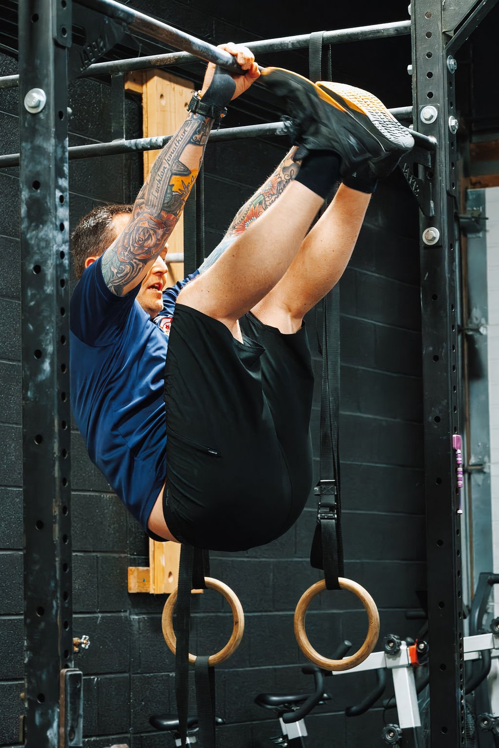 A man with tattoos wearing a blue shirt and black shorts performing an exercise on gymnastic rings attached to a pull-up bar.