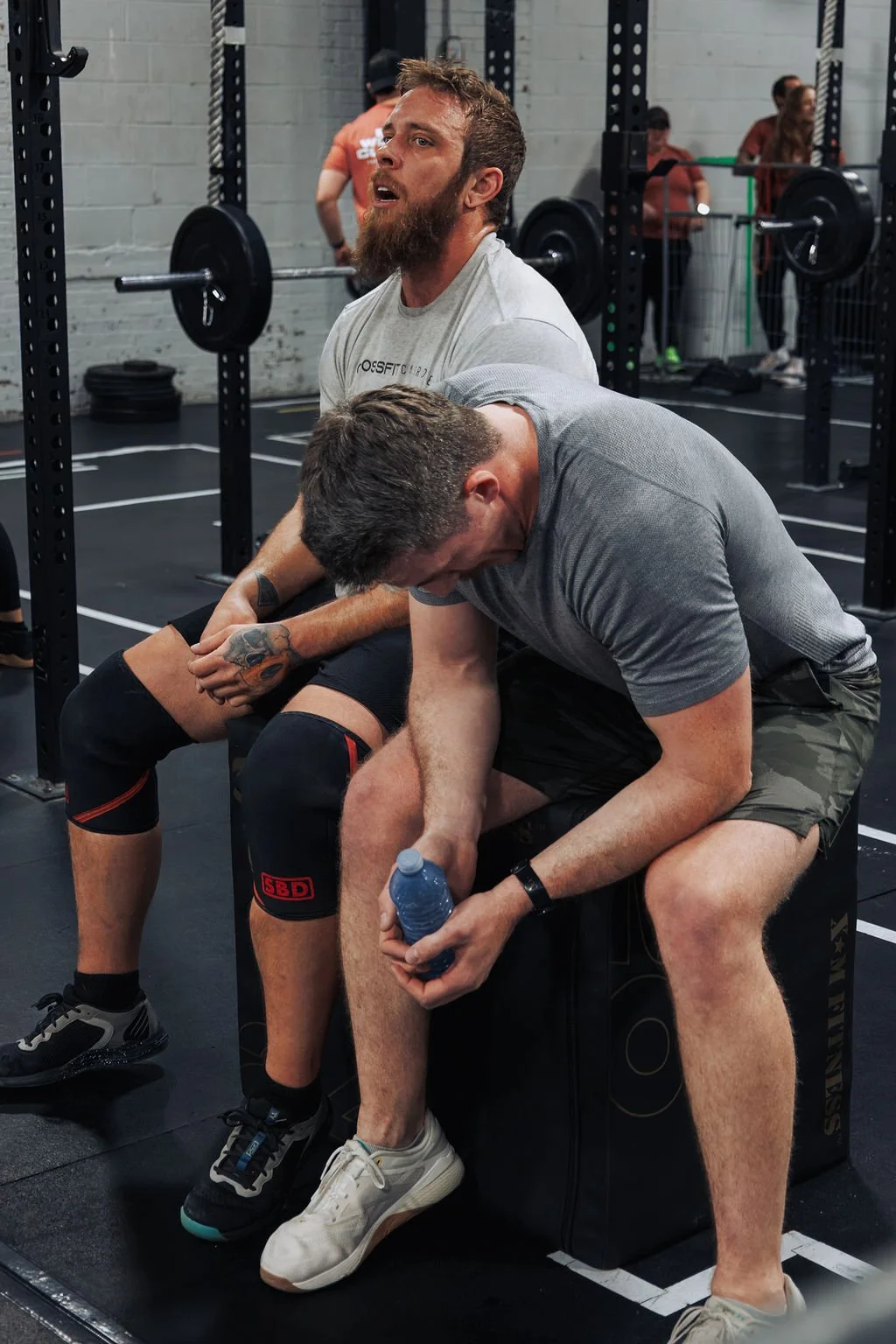 Two men resting after a workout at a gym, sitting on a black padded bench with weightlifting equipment and other people working out in the background.