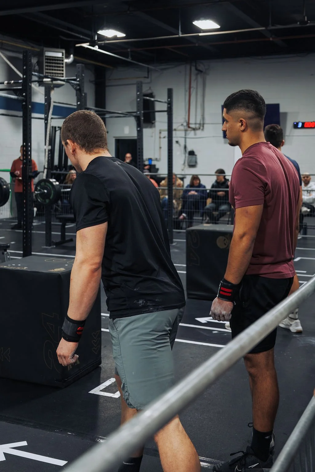 Two men preparing for a workout at a gym with weights and a metal structure, with a seated audience watching in the background.