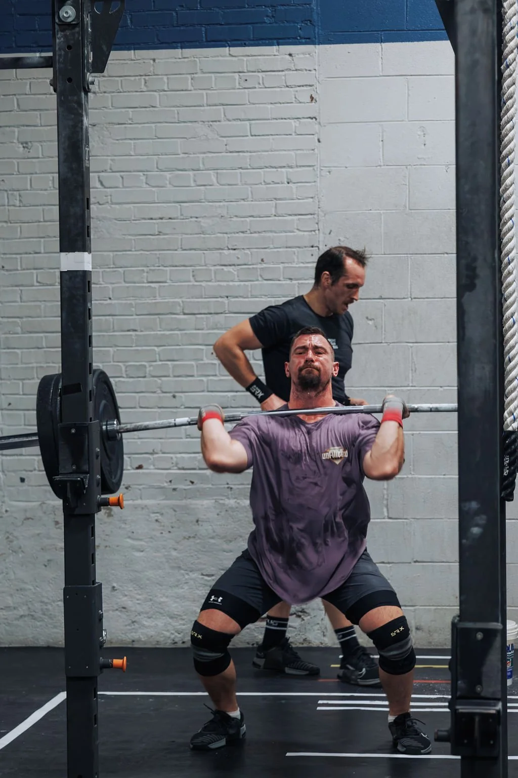 A man is lifting a barbell during a weightlifting exercise, with a coach or trainer standing behind him in a gym with brick walls.