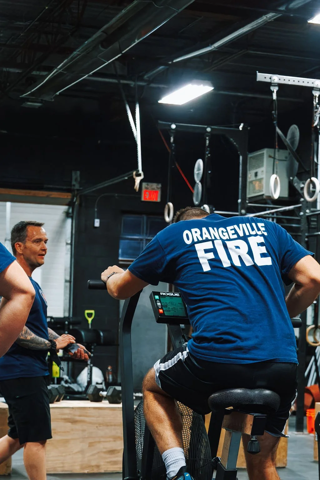 A firefighter in a blue shirt with 'Orangeville Fire' on the back is exercising on an indoor stationary bike, while another man in a blue shirt stands nearby in a gym with gymnastic rings and fitness equipment.