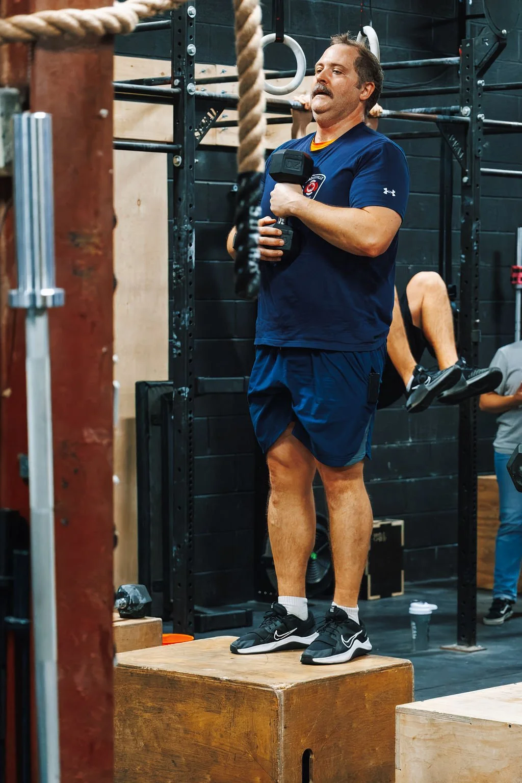A man standing on a wooden platform in a gym, holding a dumbbell with one hand, with gym equipment and another person in the background.