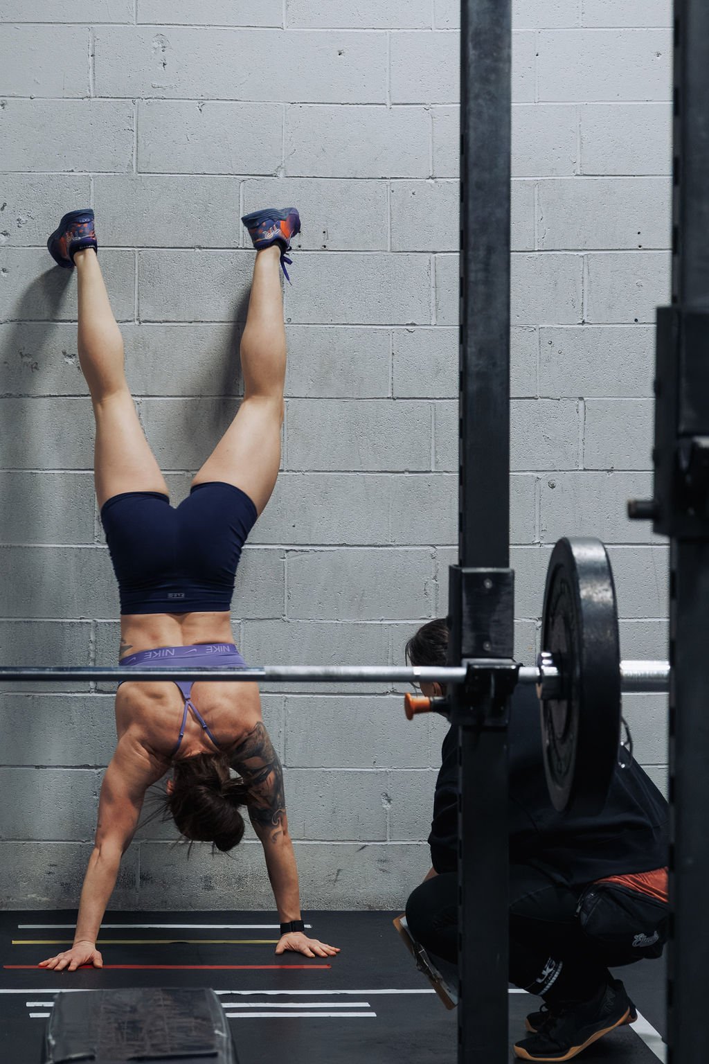 A woman is performing a handstand against a gray cinder block wall in a gym. She is wearing a purple sports bra, black shorts, and purple athletic shoes. A man is kneeling nearby, taking a photo or video of her with a phone.