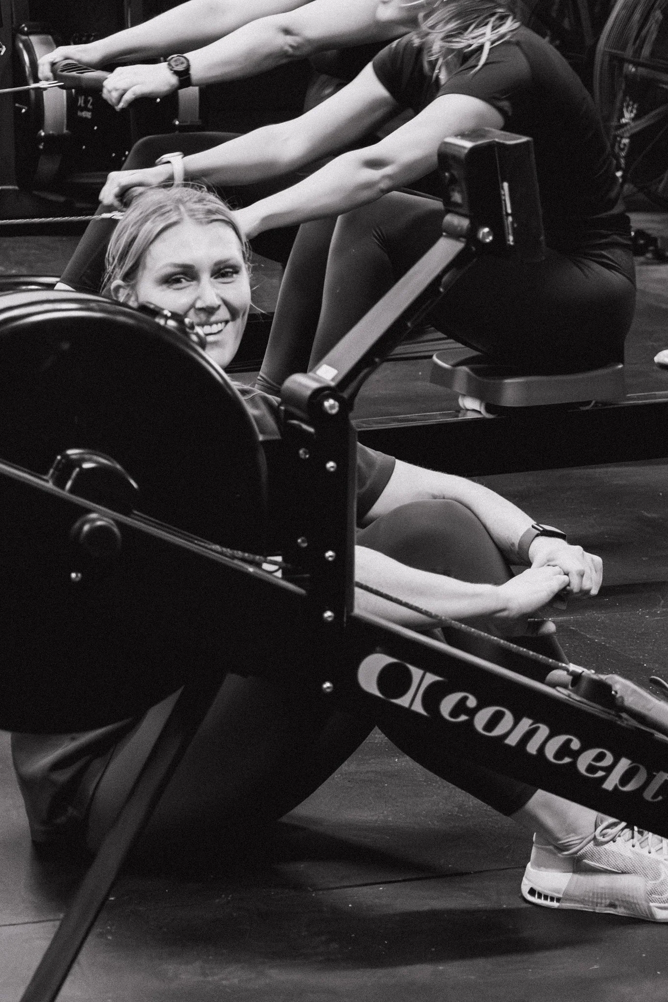 Women doing a seated rowing exercise on rowing machines in a gym, smiling at the camera.