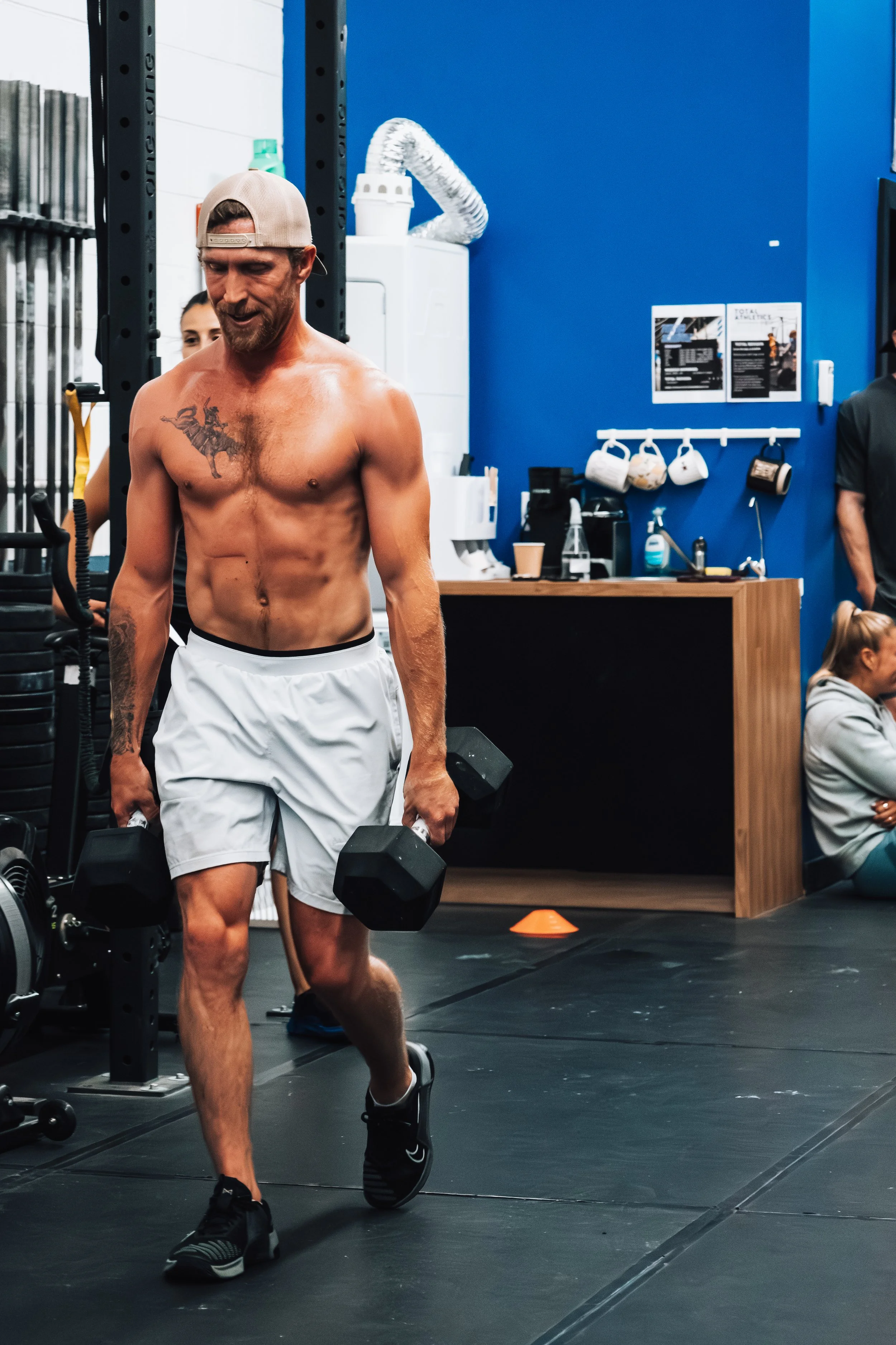 A shirtless man in a gym, wearing a baseball cap backwards, white shorts, and black sneakers, lifting dumbbells.