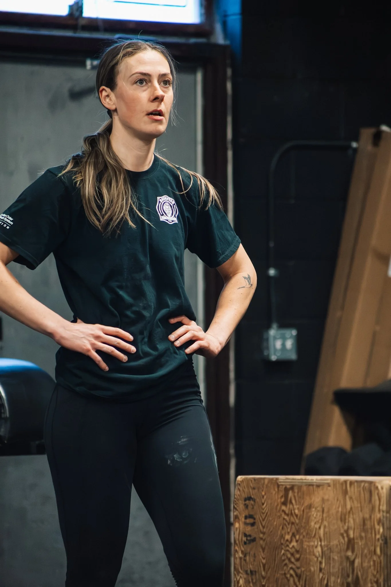 A woman with long brown hair tied back, wearing a black t-shirt with a logo on the chest and black pants, standing indoors with hands on hips, looking to the side.