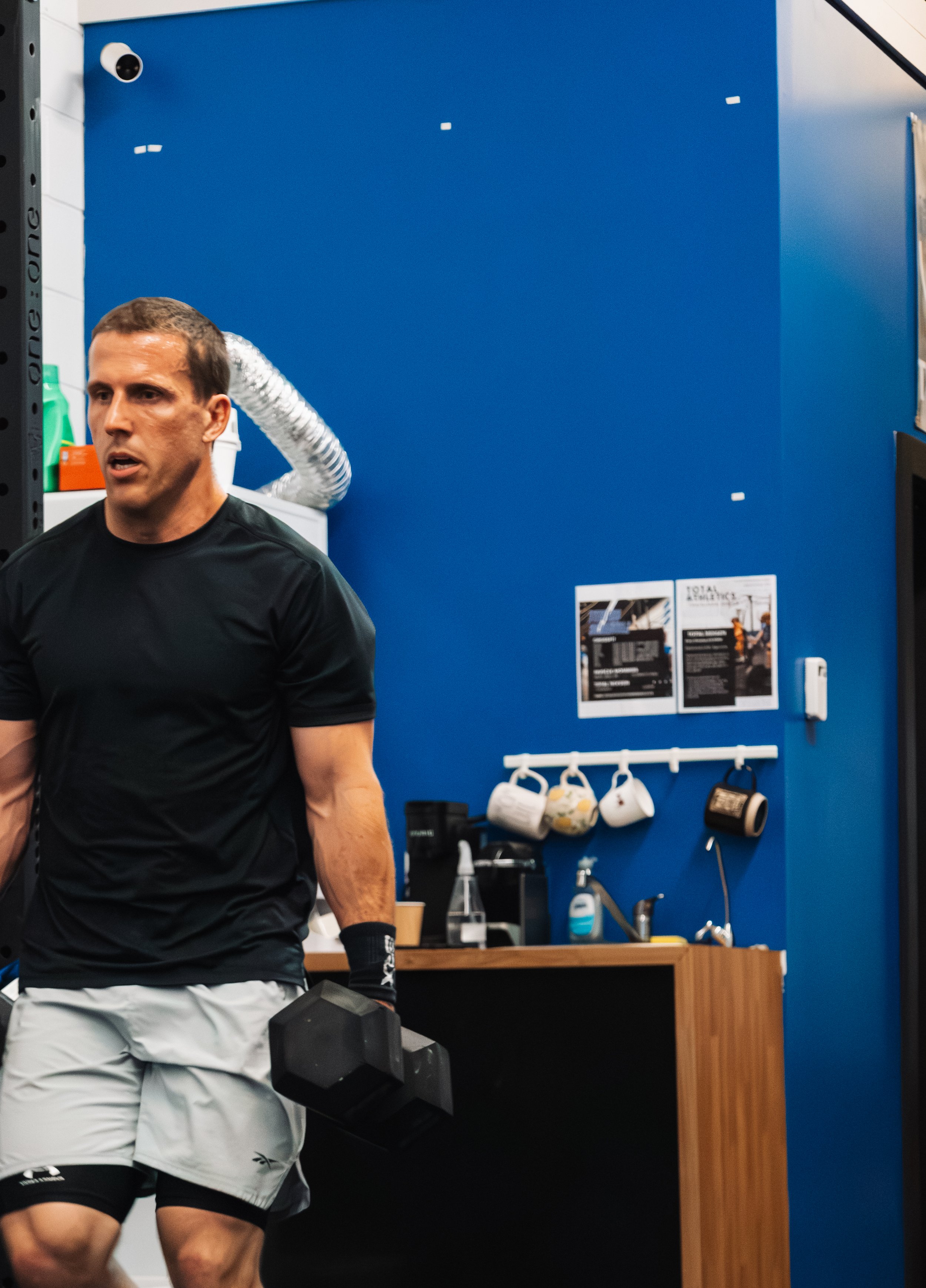 A man in workout clothes holding a dumbbell in a gym, with a blue wall and various gym equipment and posters in the background.