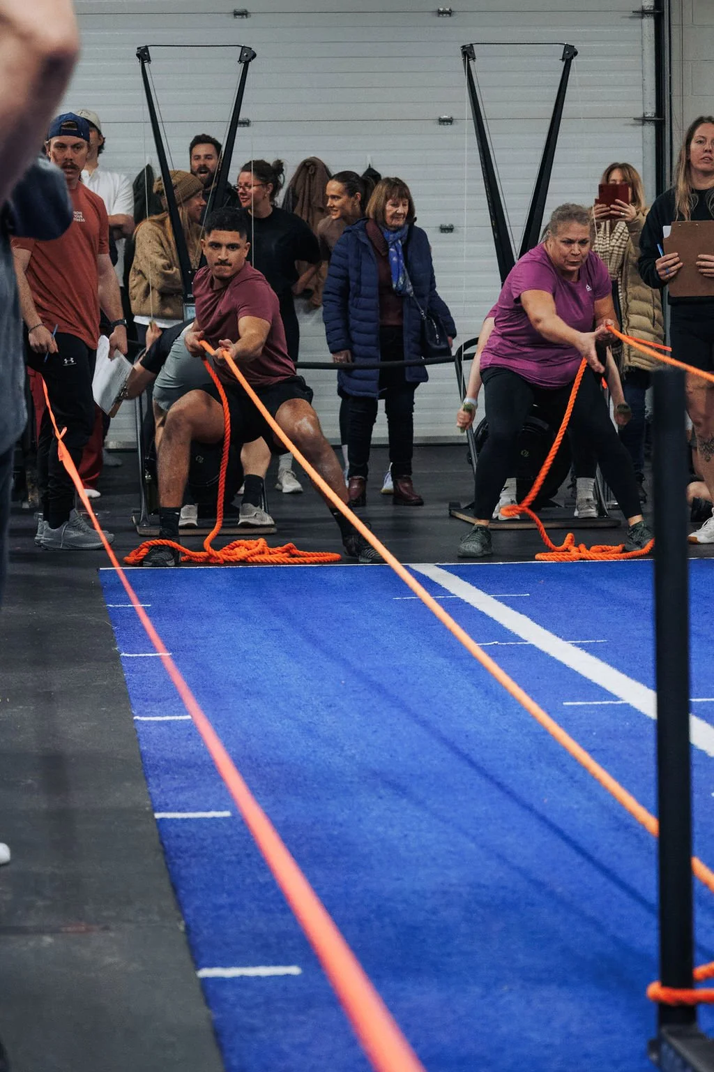 People participating in a strength challenge pulling on orange ropes in an indoor gym with a blue mat and a group of spectators watching.