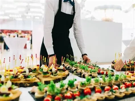 Person in uniform arranging a variety of decorated pastries and desserts on a table.