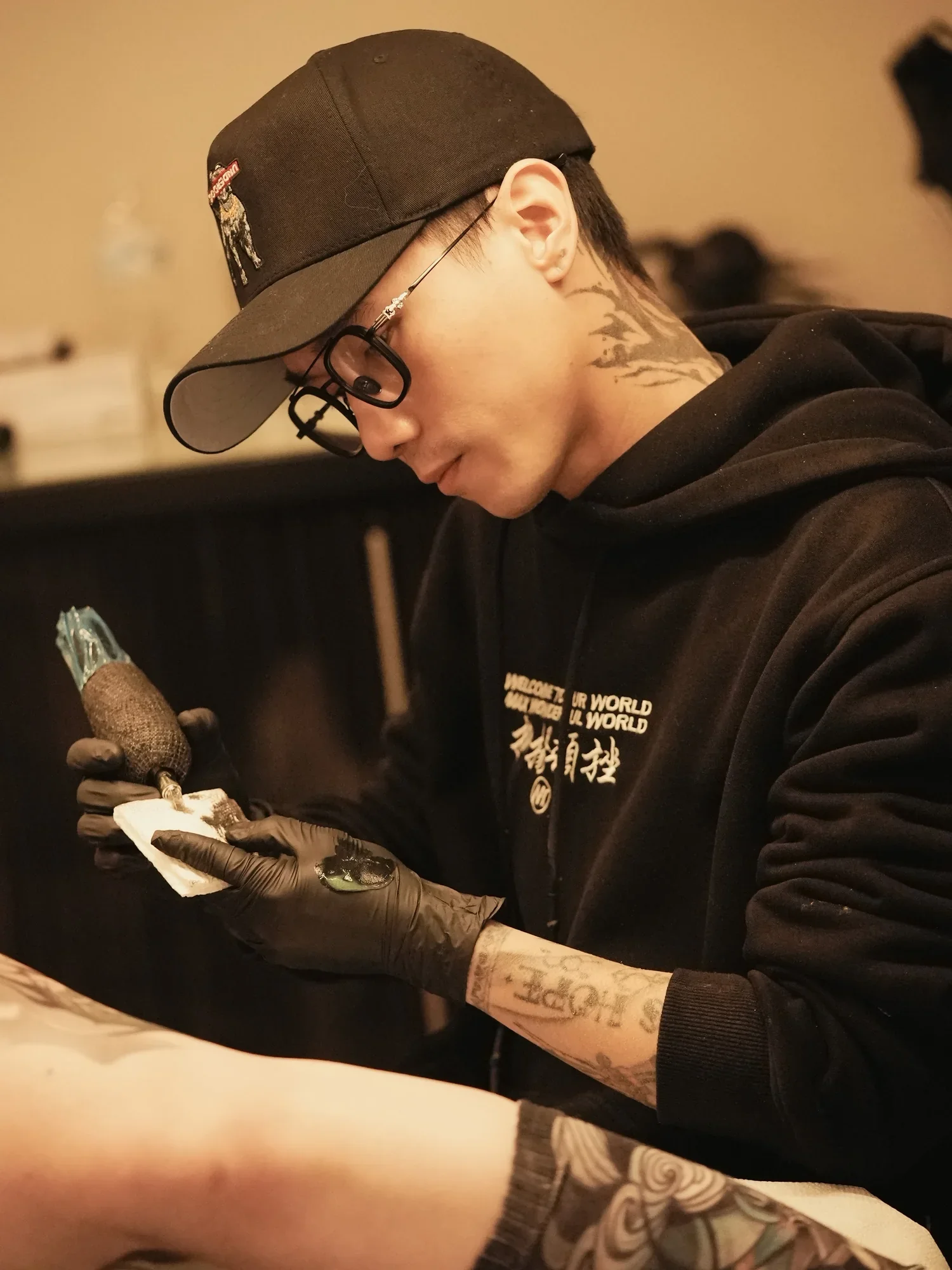 A person with tattoos on their neck and arm, wearing glasses, a black hoodie with white text, a black cap with a small logo, and black gloves, is tattooing on someone else.