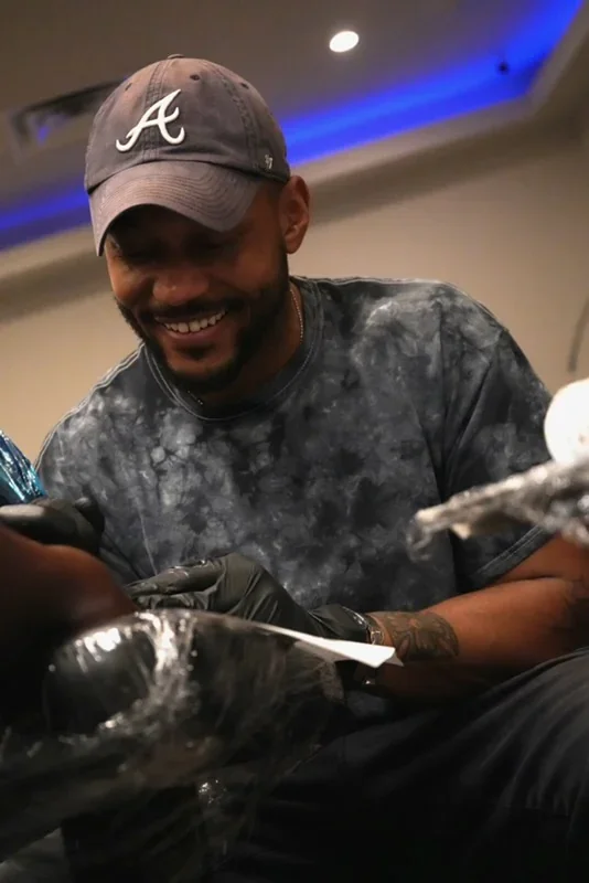 Man wearing a gray baseball cap with the Atlanta Braves logo, black gloves, smiling while looking at a phone, in a dimly lit room with blue lighting.