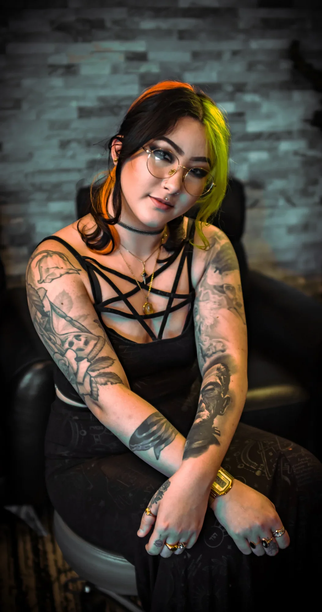 A young woman with black and yellow hair, wearing glasses, a black strappy top, and multiple jewelry pieces, sitting on a black chair against a gray stone wall background.