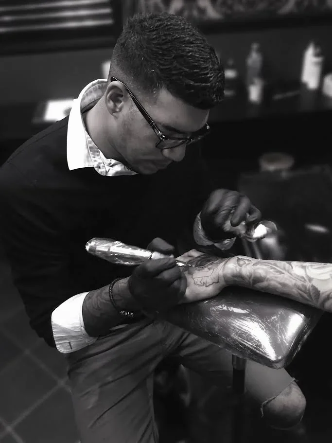 A man with glasses, short dark hair, and tattoos on his arm is tattooing a person's hand in a tattoo studio, using a tattoo machine.