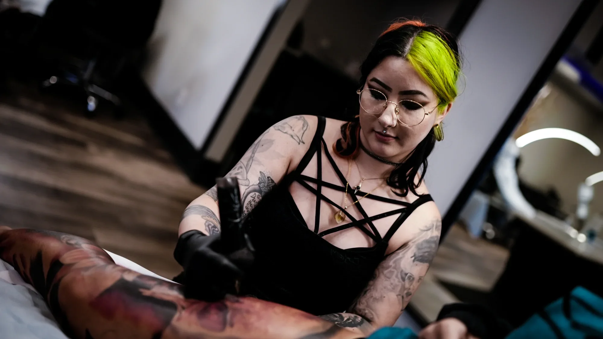 A tattooed woman with black and neon green hair and glasses is tattooing a person's leg. She is wearing a black strappy top and has tattoos on her arms. The setting appears to be a tattoo studio.