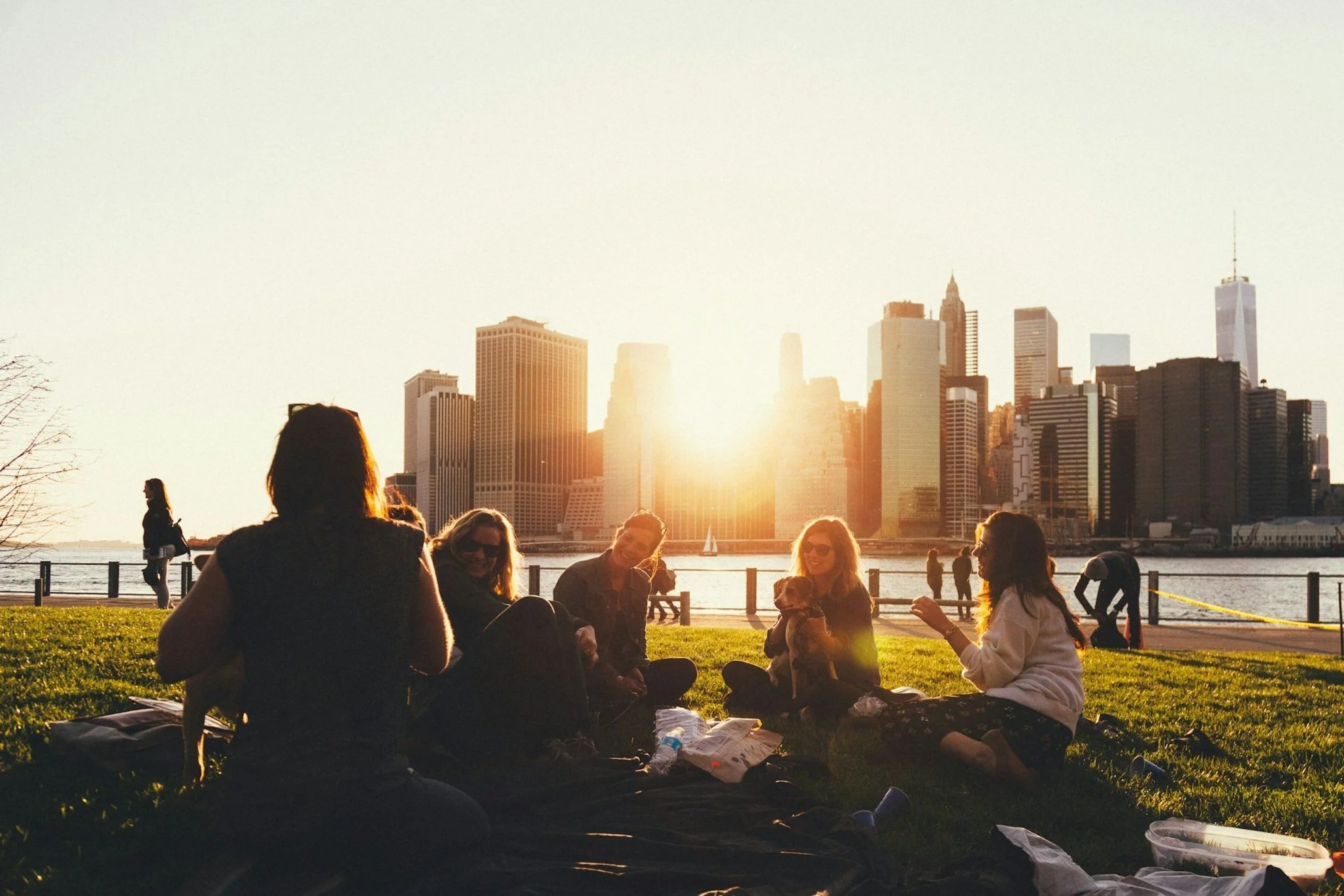 Group of people having a picnic in a park with a city skyline at sunset.
