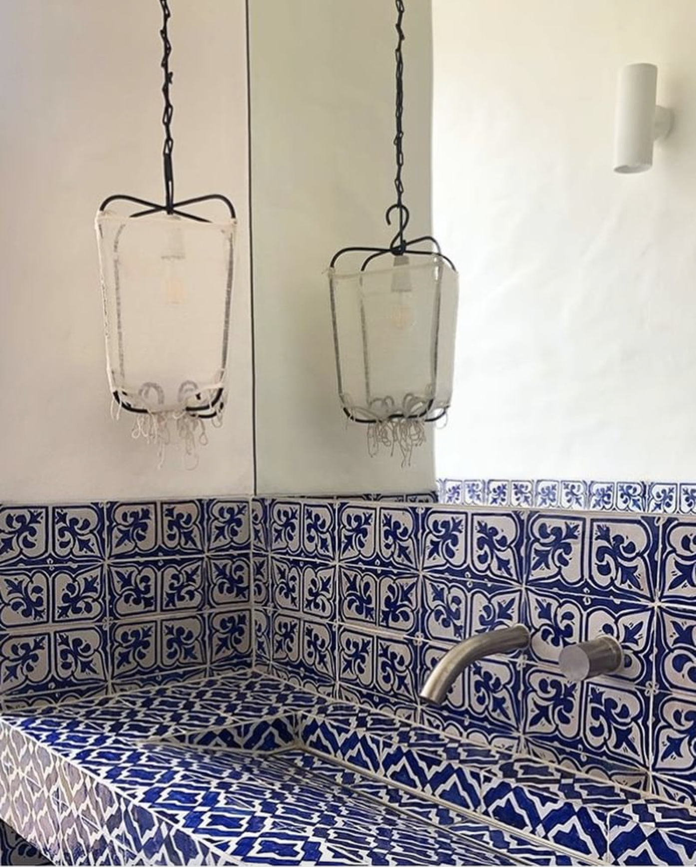 A bathroom with a tiled blue and white designed countertop and wall. Two hanging lantern-style light fixtures are reflected in a mirror. A faucet is mounted on the tiled wall.
www.gardenlampcollection.com