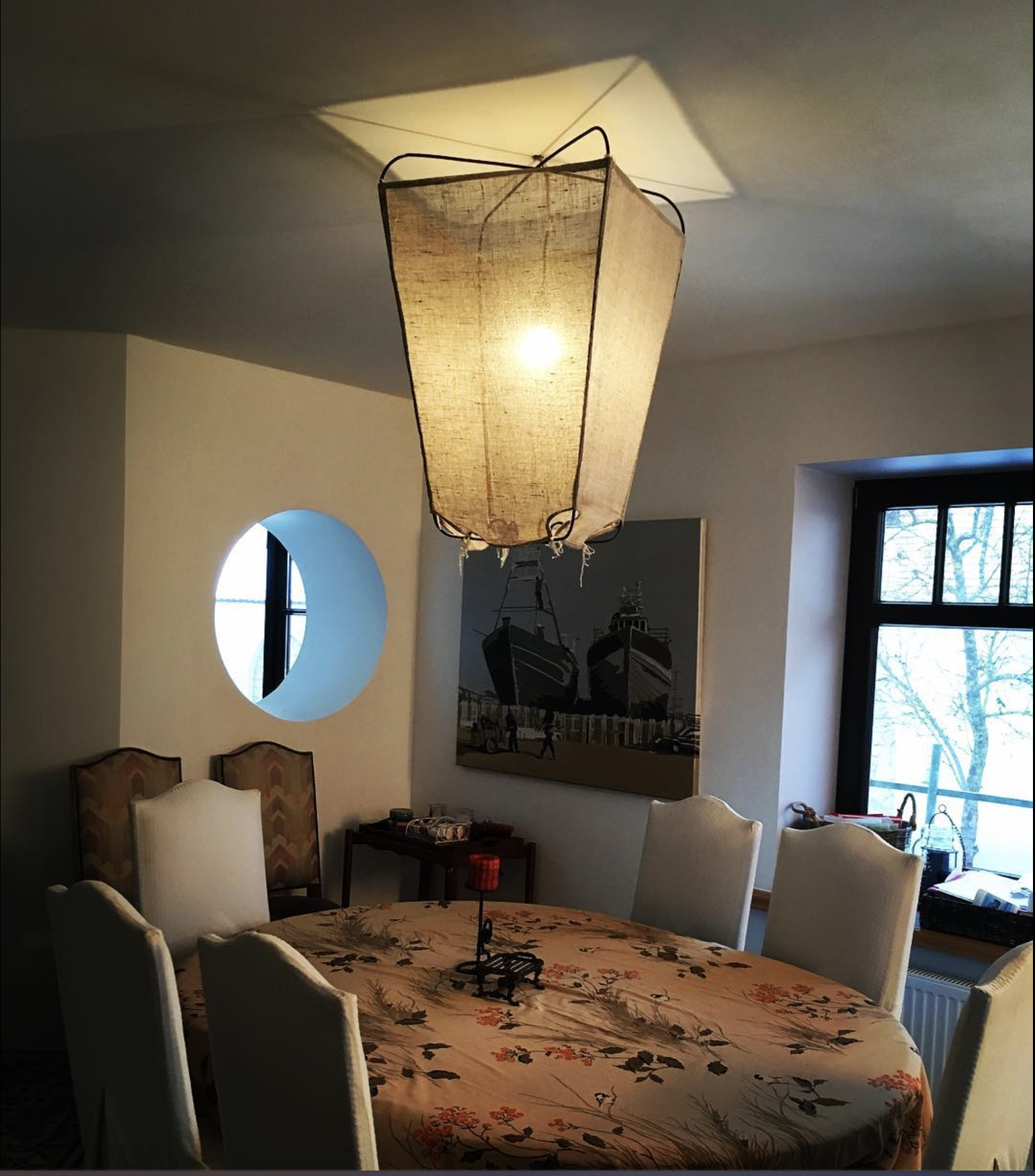 A dining room with a round table draped with a floral tablecloth, six cream-colored chairs around it, a rectangular chandelier hanging above, and a window allowing natural light in. www.gardenlampcollection.com