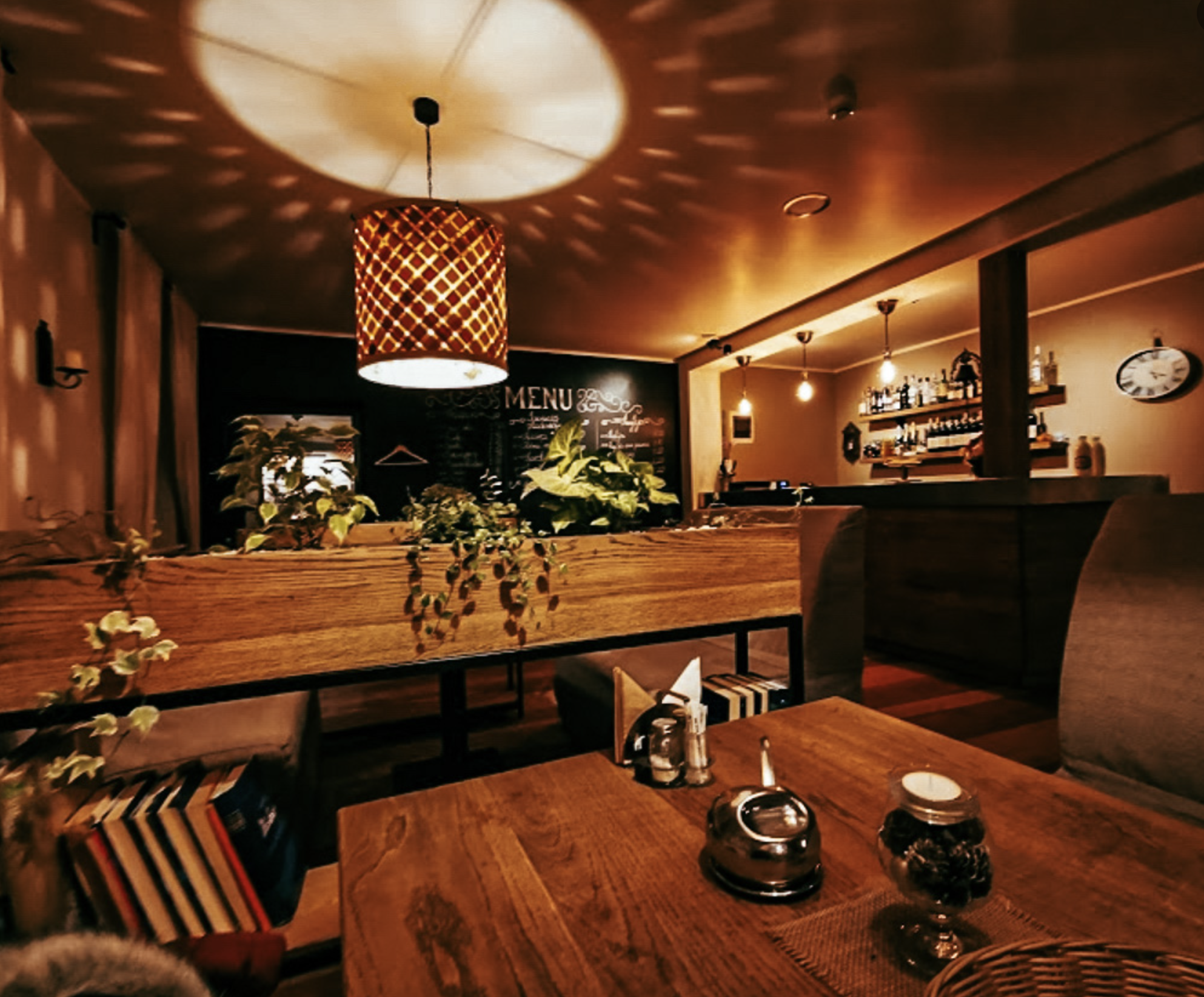 Cozy dimly-lit restaurant interior with wooden tables, plants, decorative hanging lamps, a chalkboard menu, and a bar area in the background.
www.gardenlampcollection.com