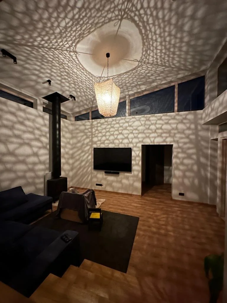 Living room with a large flat-screen TV, a black sofa, a wooden floor, a patterned paper lantern lighting fixture casting shadows on the ceiling and wall, and a black wood stove in the corner.
www.gardenlampcollection.com