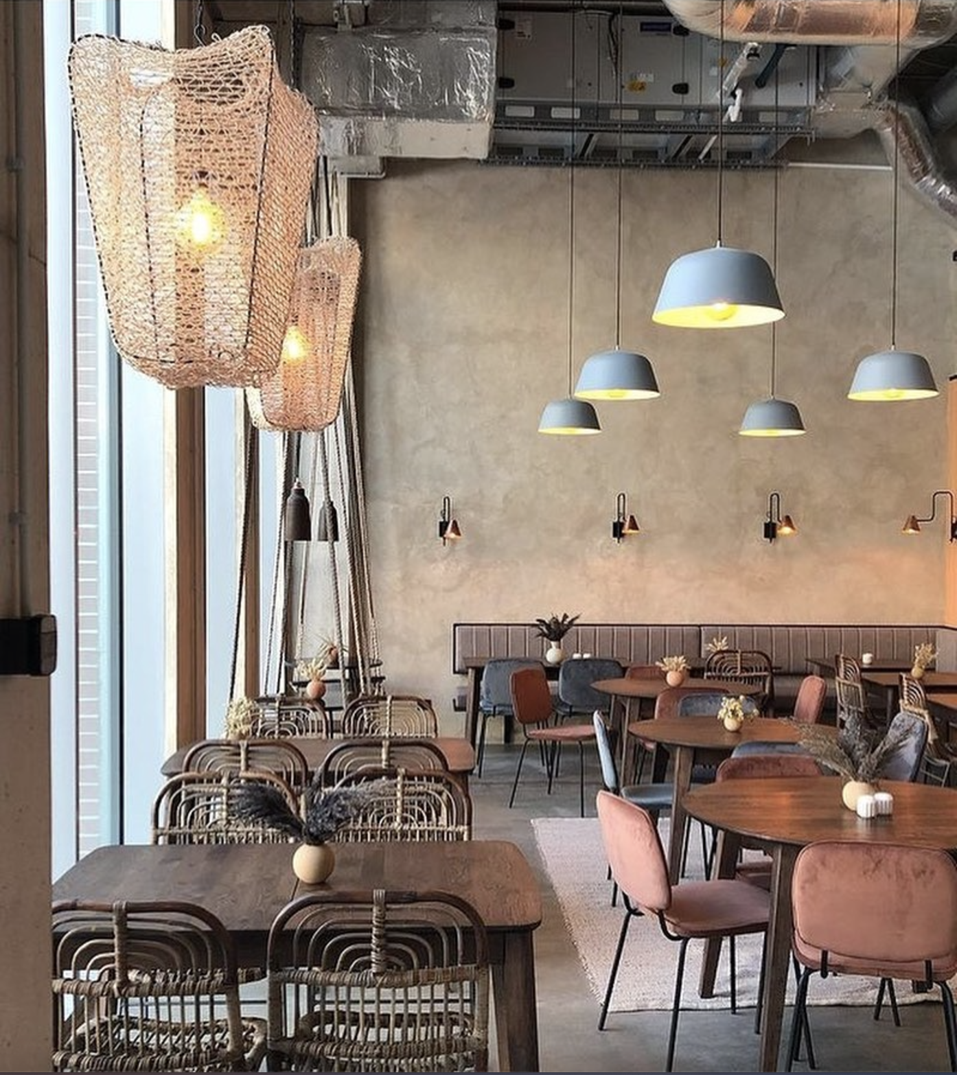 Modern cafe with pendant and wall lighting, wooden tables, pink and gray upholstered chairs, and small vases with dried flowers on each table, next to large front windows.
www.gardenlampcollection.com