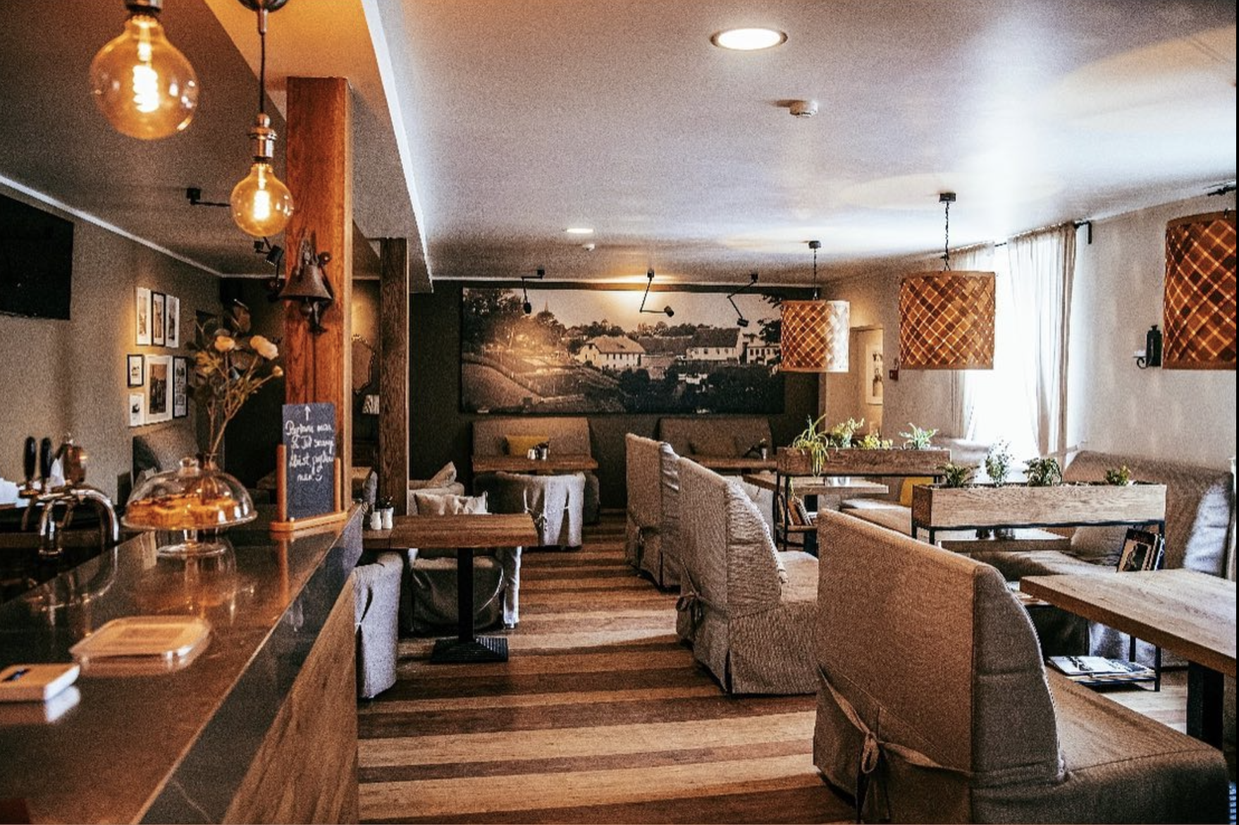 A cozy restaurant interior with wooden floors, beige upholstered chairs, and window curtains. Decorative hanging lamps and wall art are visible.
www.gardenlampcollection.com