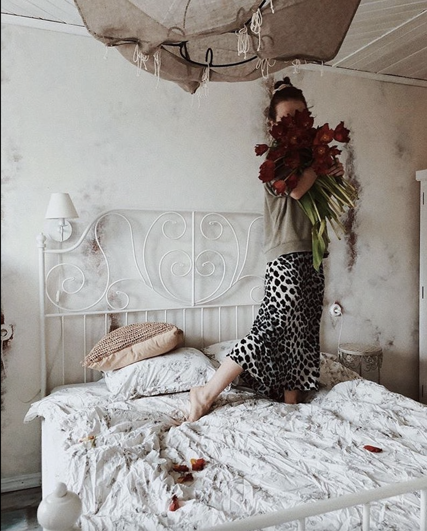 A woman standing on a bed with a rumpled white comforter, holding a large bouquet of red flowers, in a bedroom with a white metal bed frame, patterned pillow, a small wall-mounted lamp, and a textured wall with peeling paint.