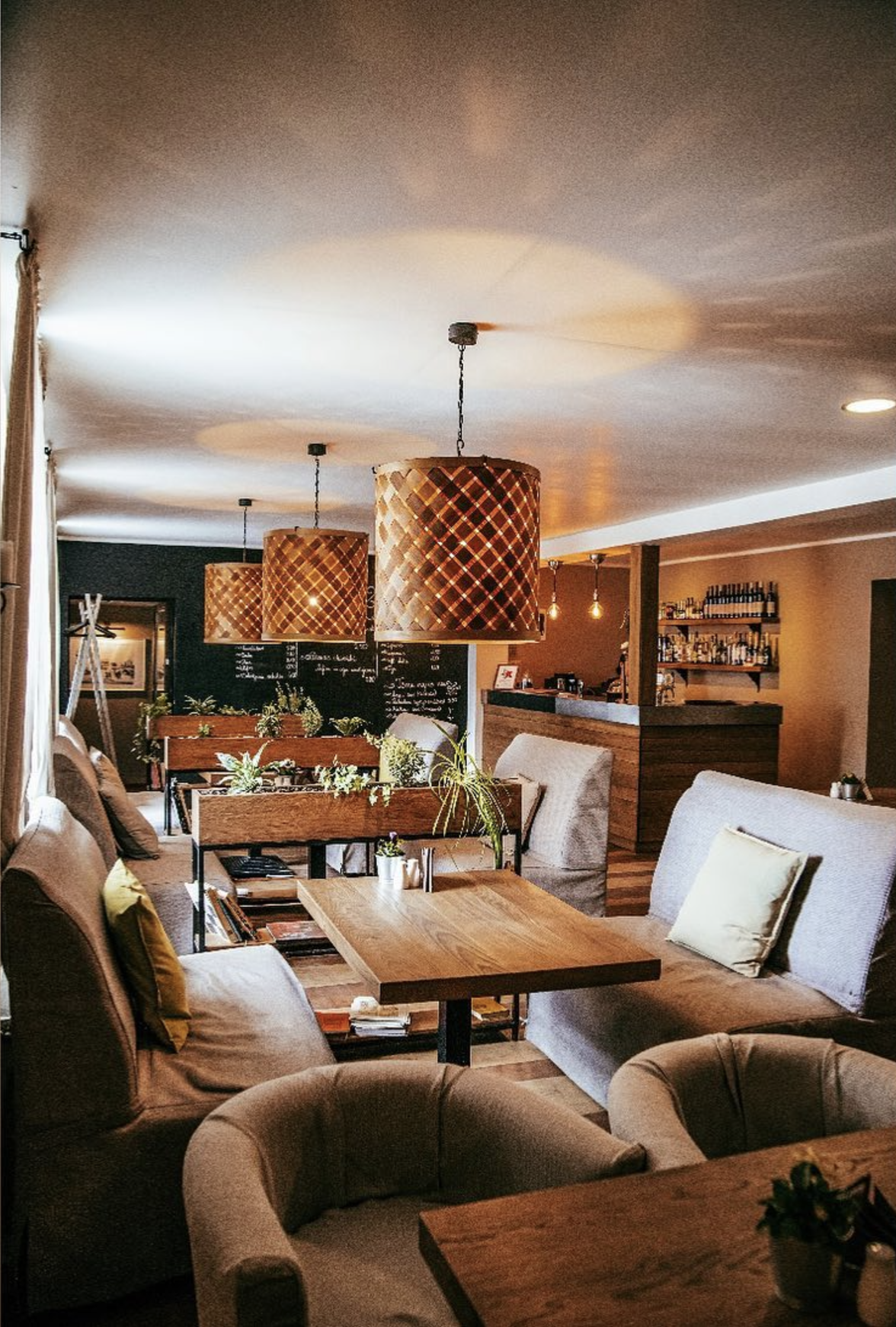 Interior of a cozy cafe with wooden tables, beige upholstered chairs, hanging woven pendant lights, and plants, with a bar in the background.
www.gardenlampcollection.com