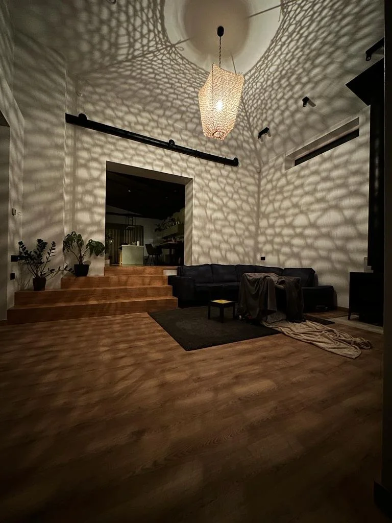 Living room with wooden floors, a black sectional sofa with a throw blanket, a small black coffee table, and a black rug. A hanging lampshade creates intricate light and shadow patterns on the wall and ceiling.
www.gardenlampcollection.com