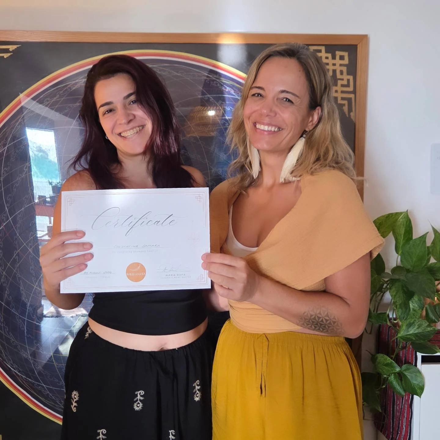 Two smiling women holding a certificate together, standing in front of a space-themed poster and a potted plant.