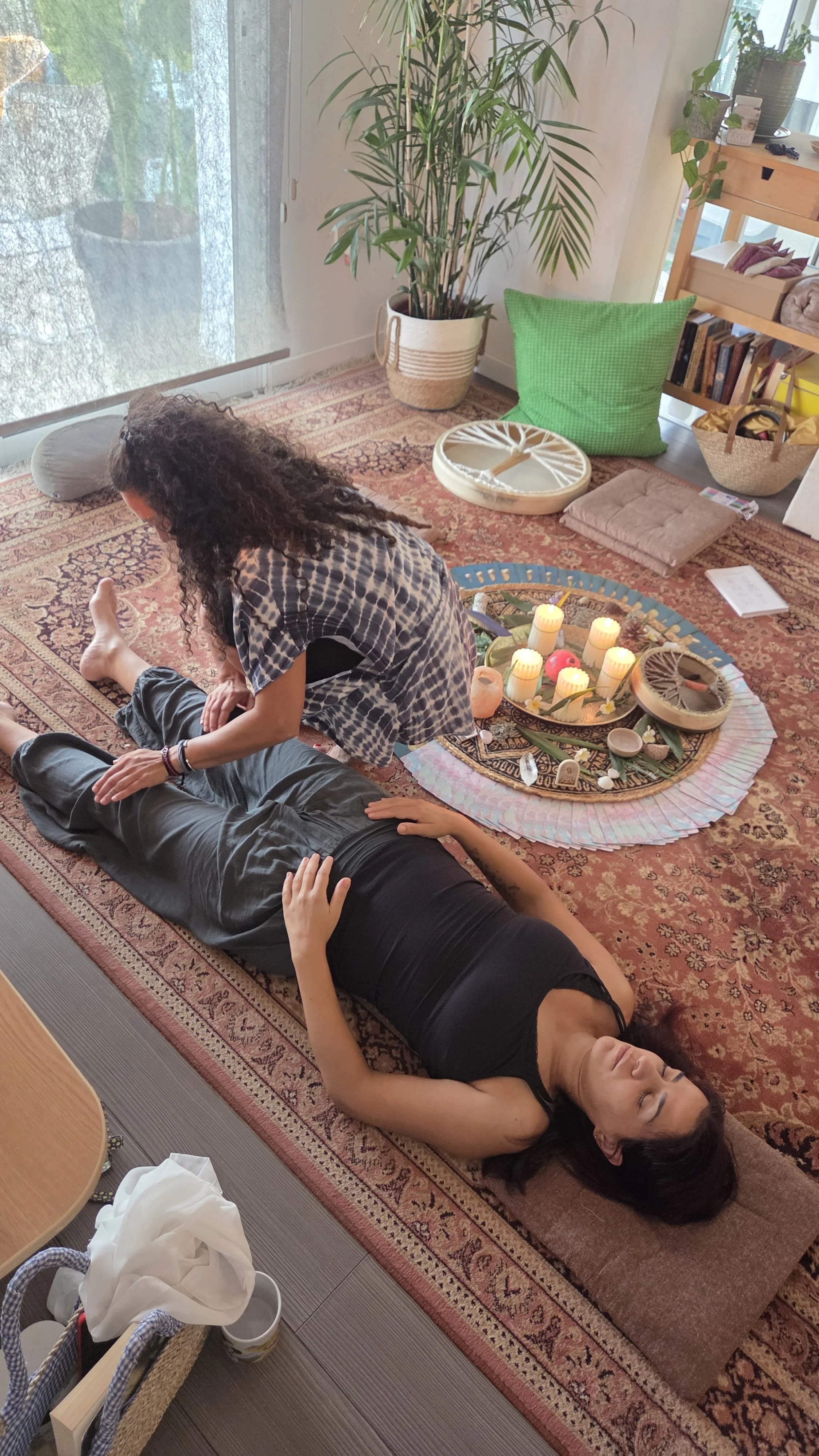 A woman lying on a carpet receiving a massage from another woman. The setting appears to be a cozy indoor space with plants, books, and decorated with candles and ornaments on a round tray.