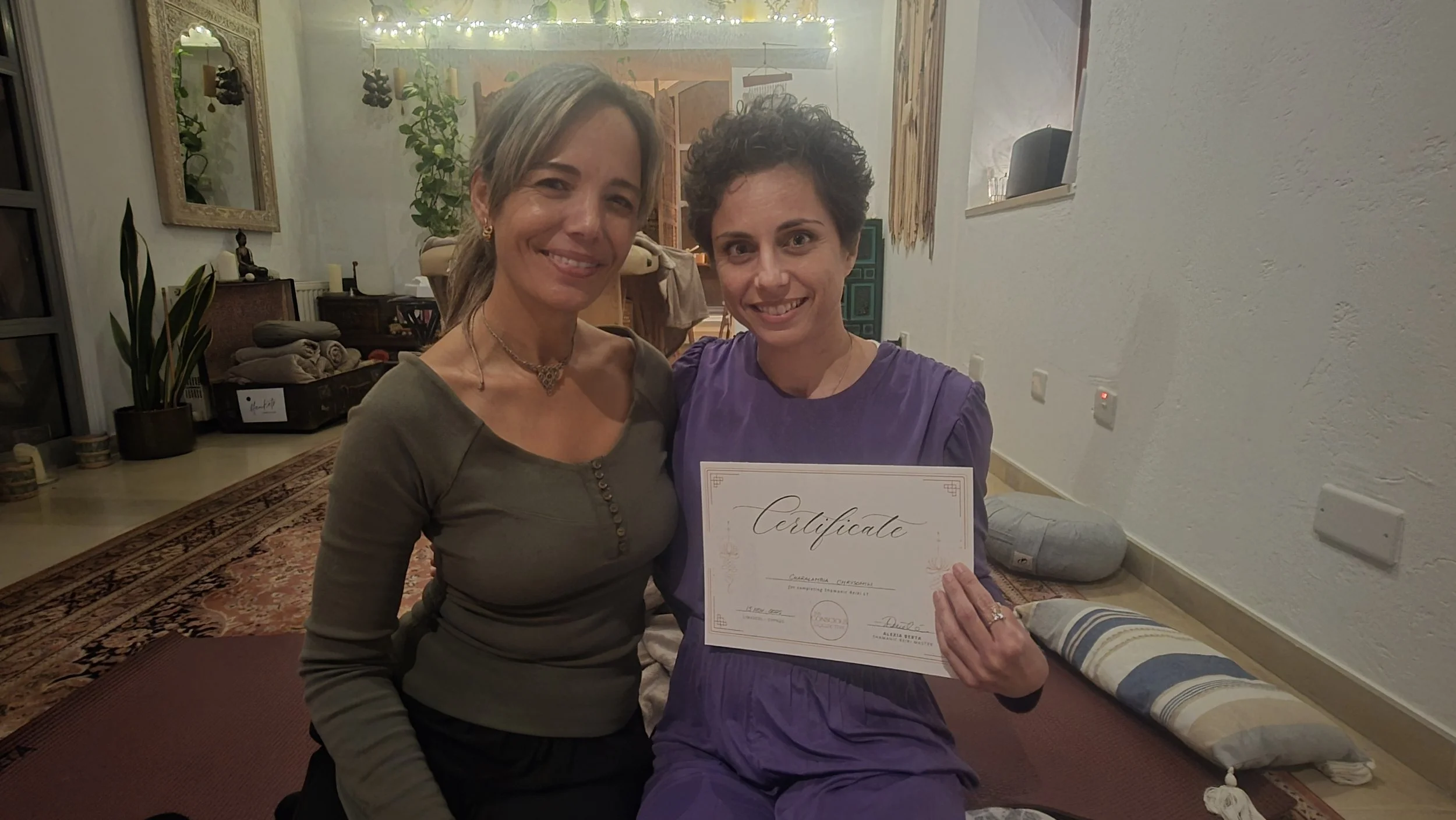 Two women smiling, one holding a certificate, celebrating indoors with warm lighting, plants, and decorative items in the background.
