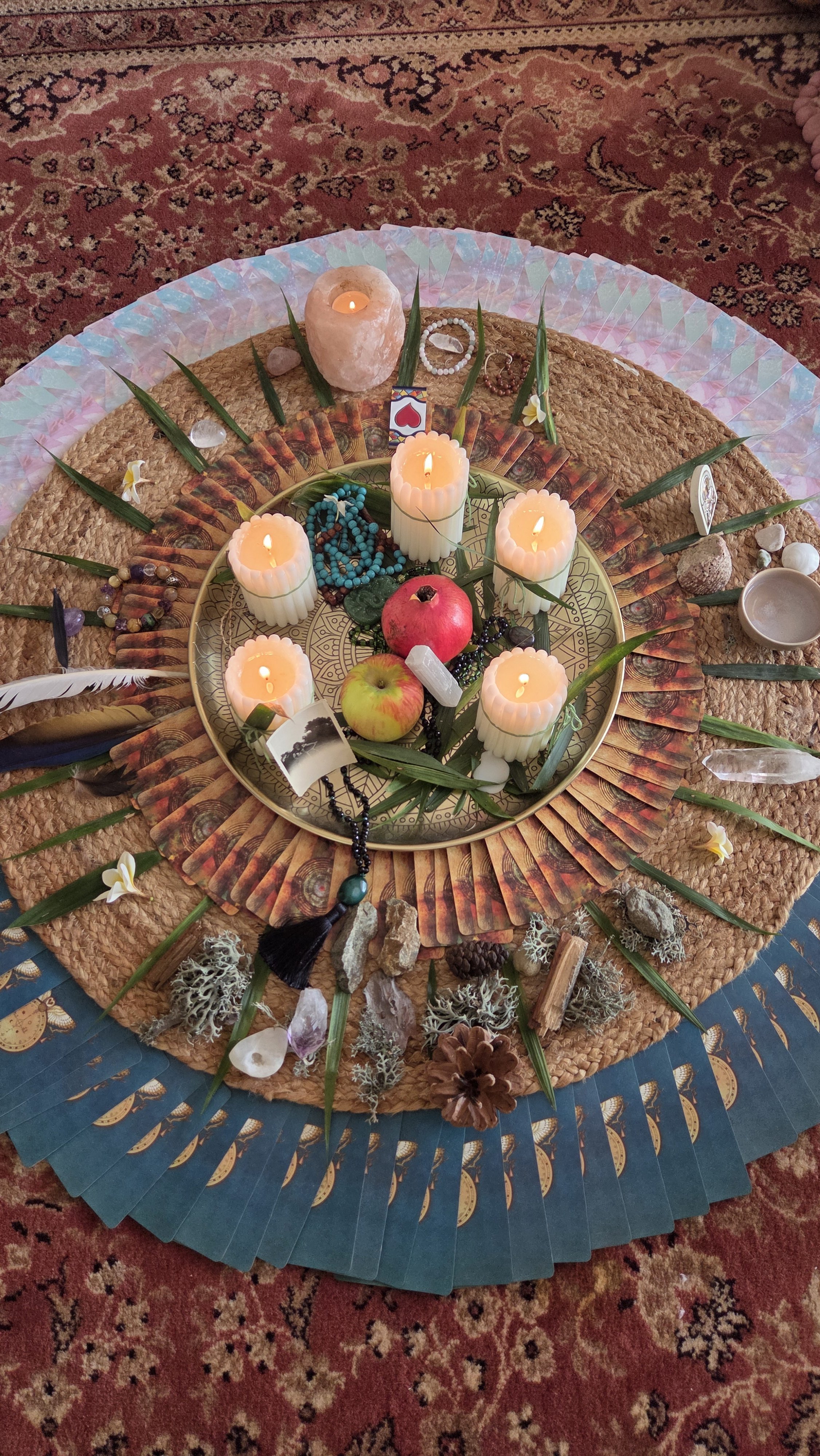 A circular arrangement on a woven mat with candles, roses, apples, crystals, beads, and natural objects, surrounded by a colorful fan, on a patterned carpet.