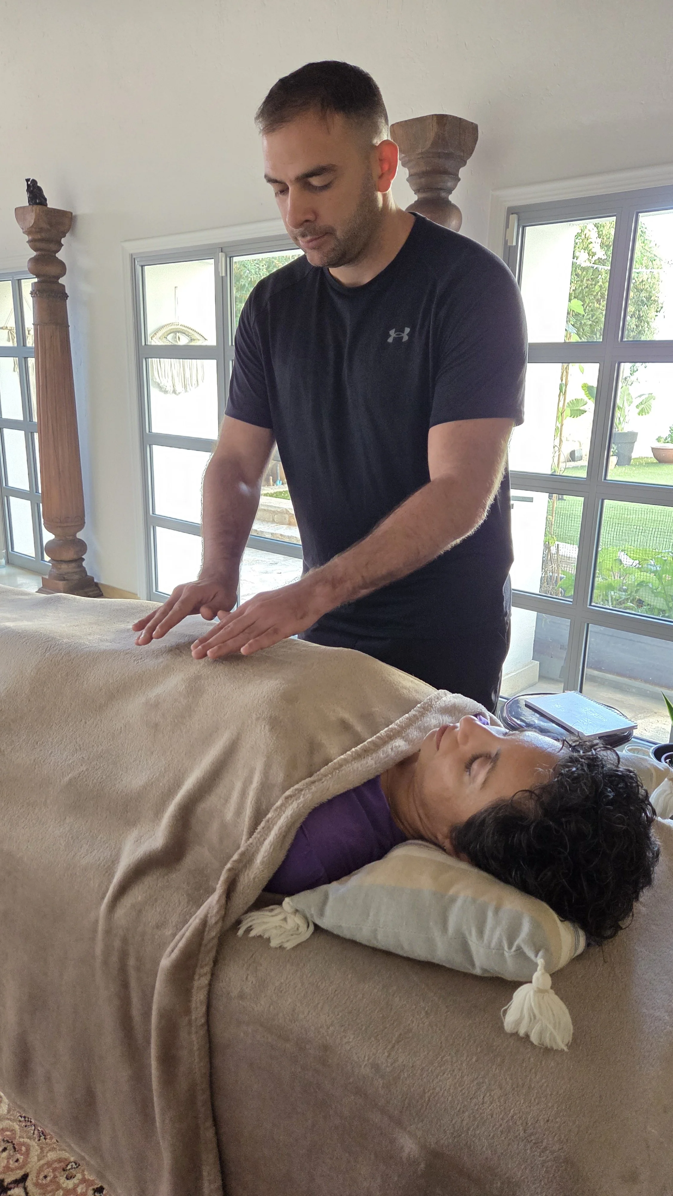 A man practicing massage or physical therapy on a woman lying on a massage table in a bright room with large windows and garden view.