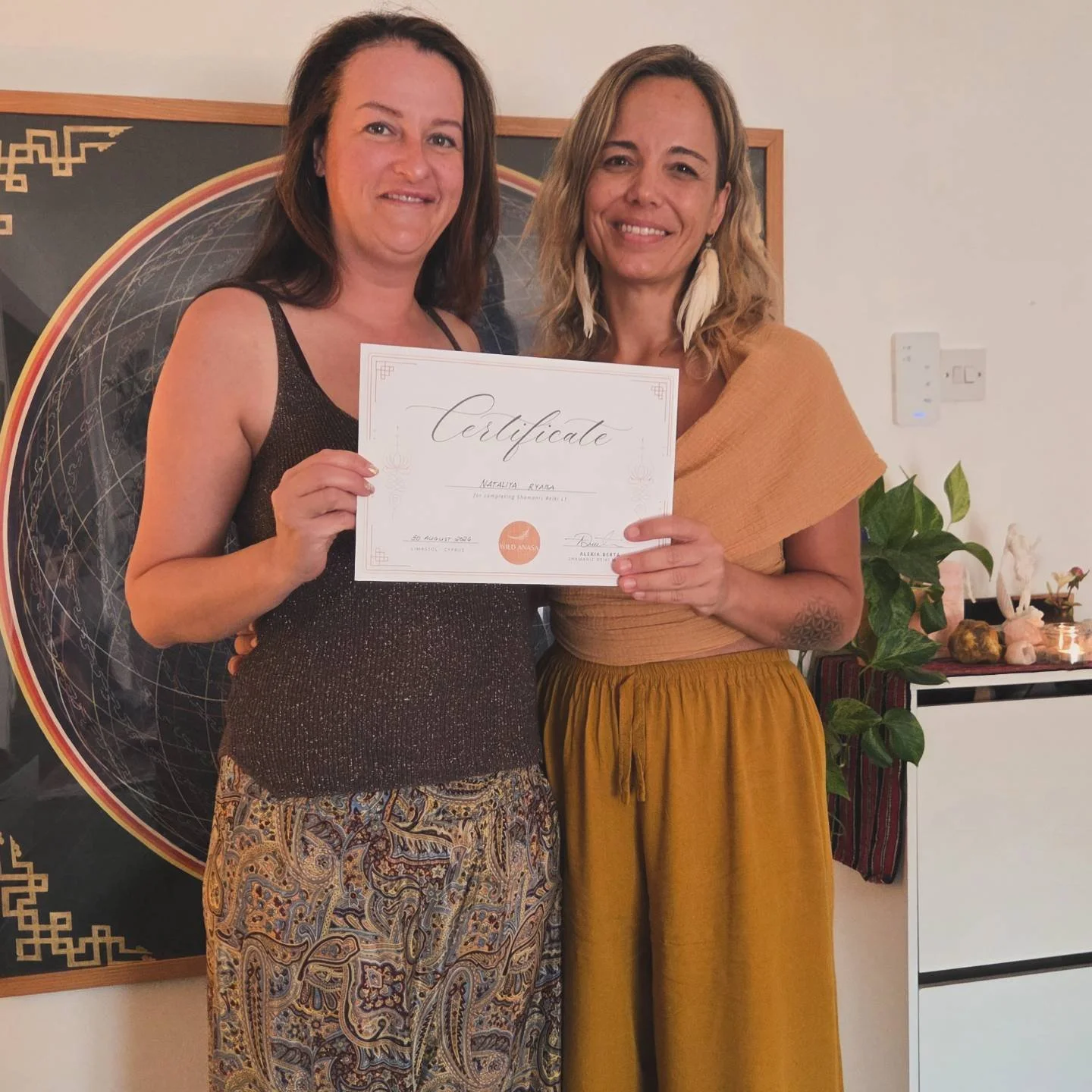 Two women smiling, holding a certificate, standing indoors against a decorative backdrop, with plants and ornaments on a nearby table.