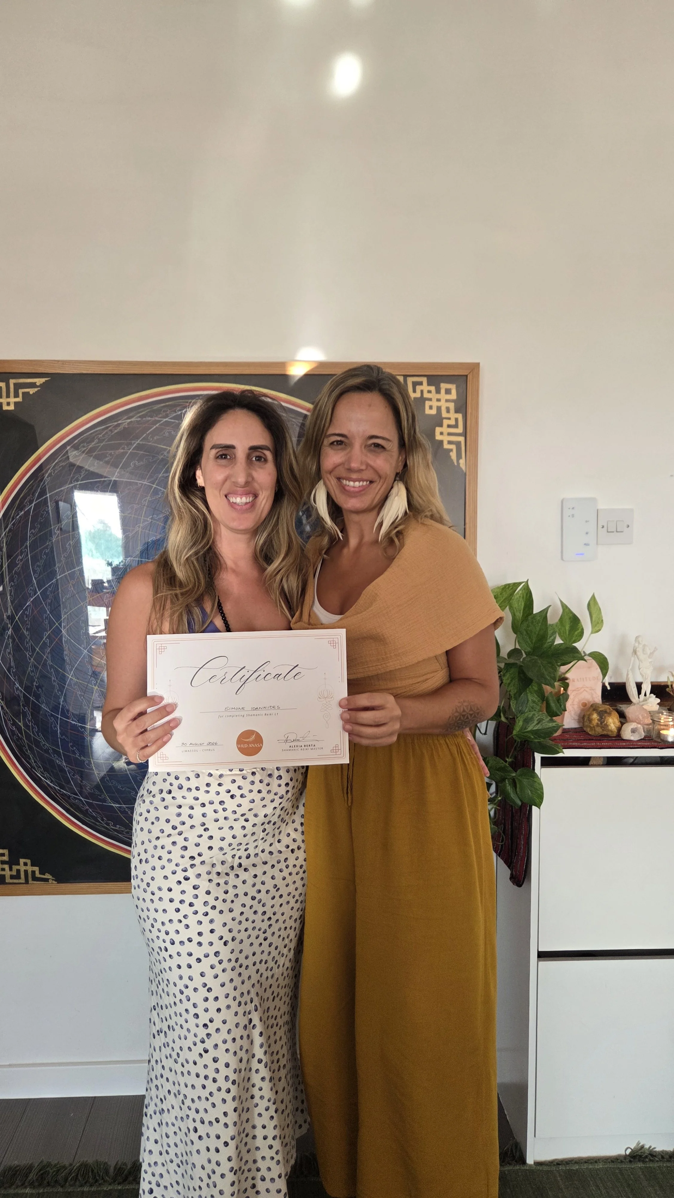 Two women standing side by side, smiling, with one holding a certificate. They are indoors with a decorative background, plants, and crystals on a table in the background.