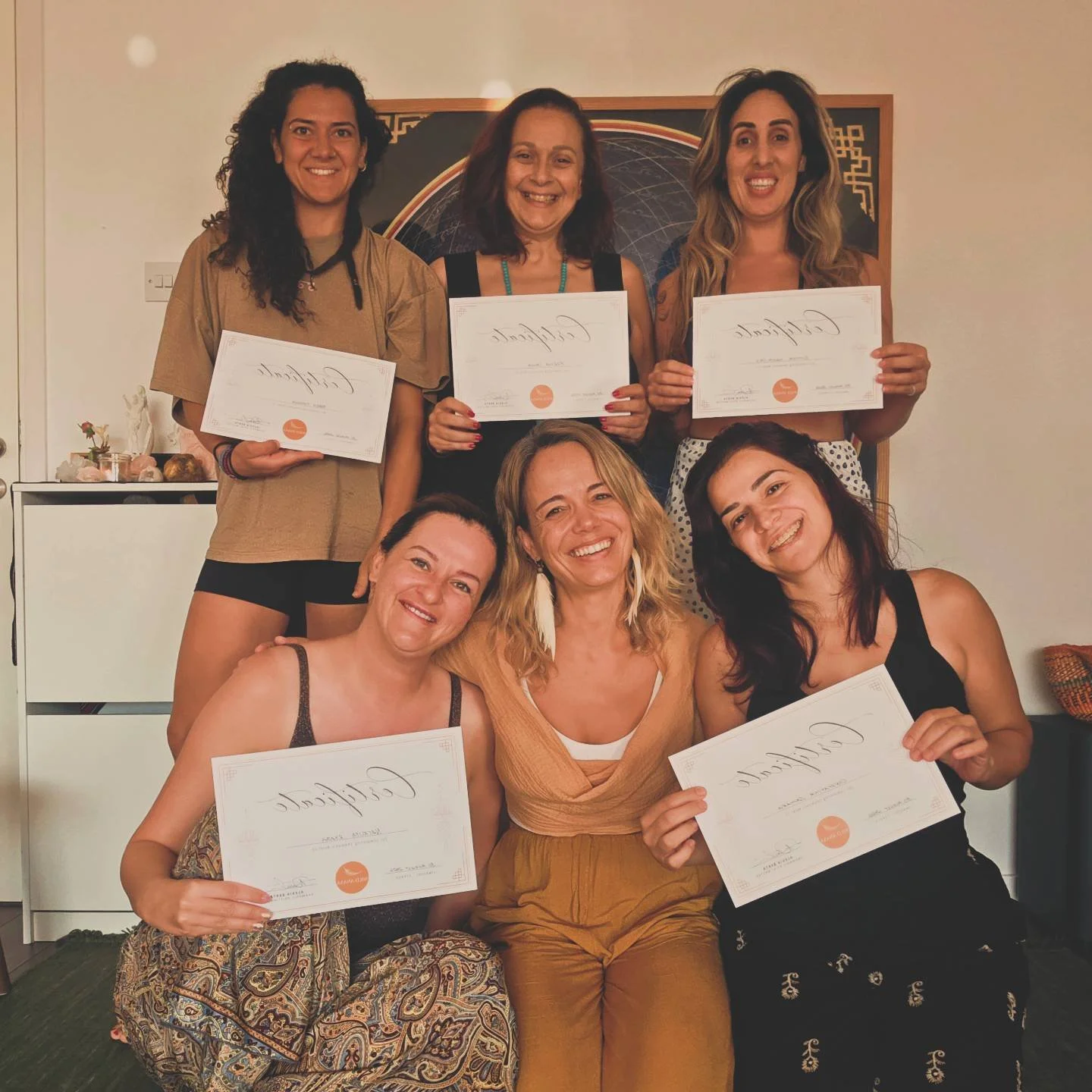 Six women posing with certificates in a room, smiling, with a decorative wall hanging in the background.