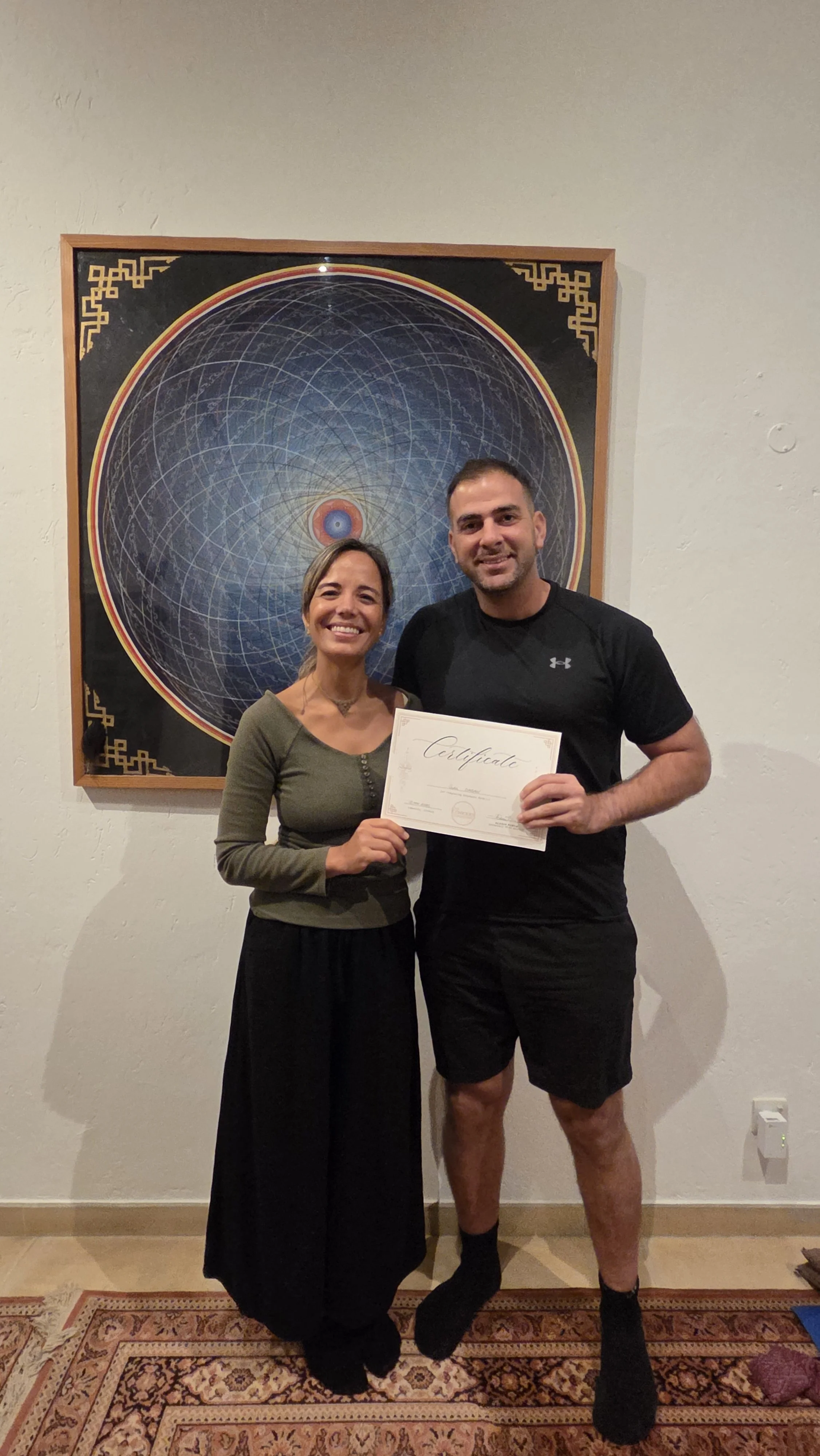 A woman and man standing indoors in front of a framed picture on a white wall. The woman is holding a certificate and smiling, wearing a green top and black skirt. The man is wearing a black Under Armour t-shirt and shorts, smiling, with his arm around the woman.